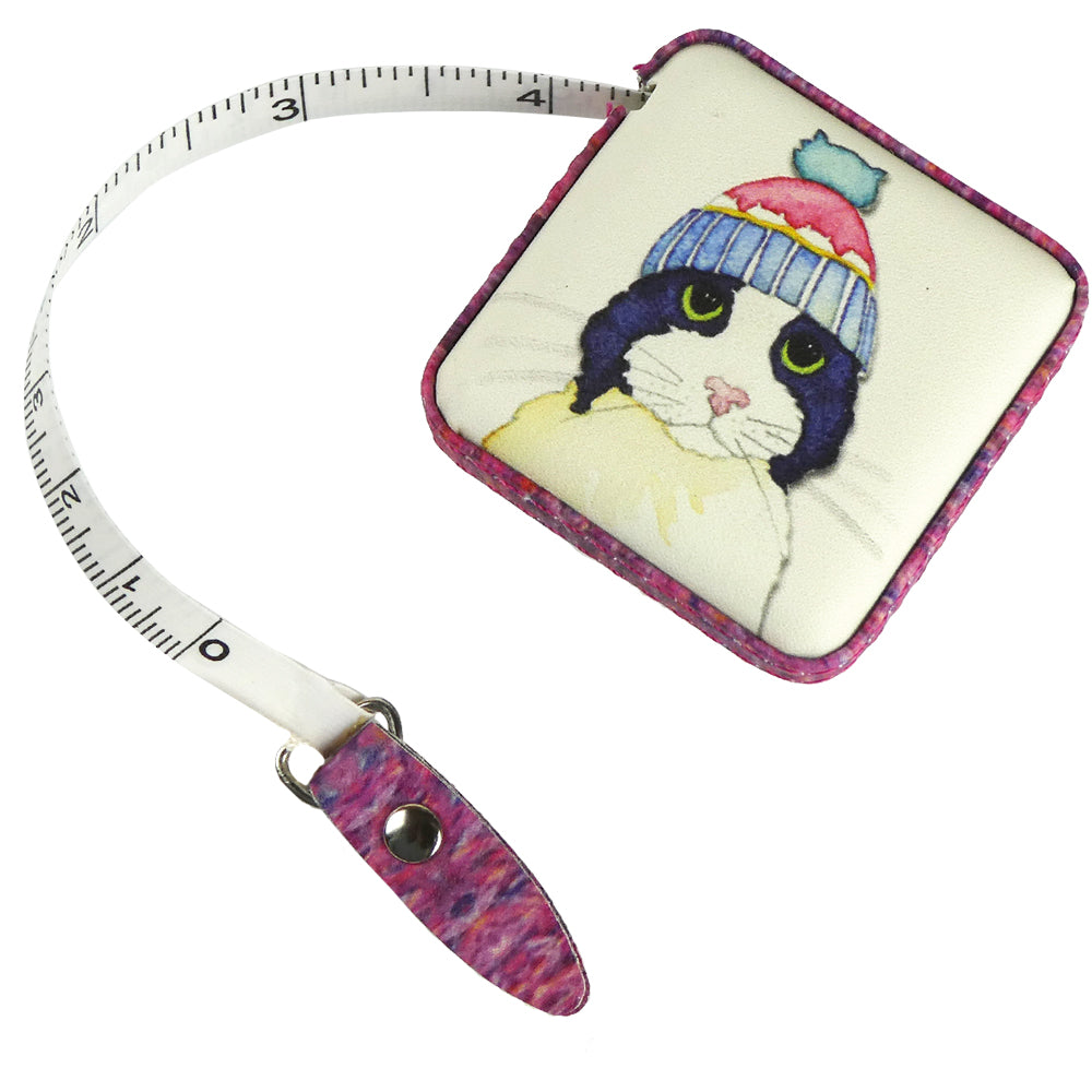 Quadratisches Maßband mit ausgezogener Inch-Skala. Aquarell-Illustration von Emma Ball: schwarz-weiße Katze mit großen grünen Augen in rosa-blauer Strickmütze und gelbem Schal.