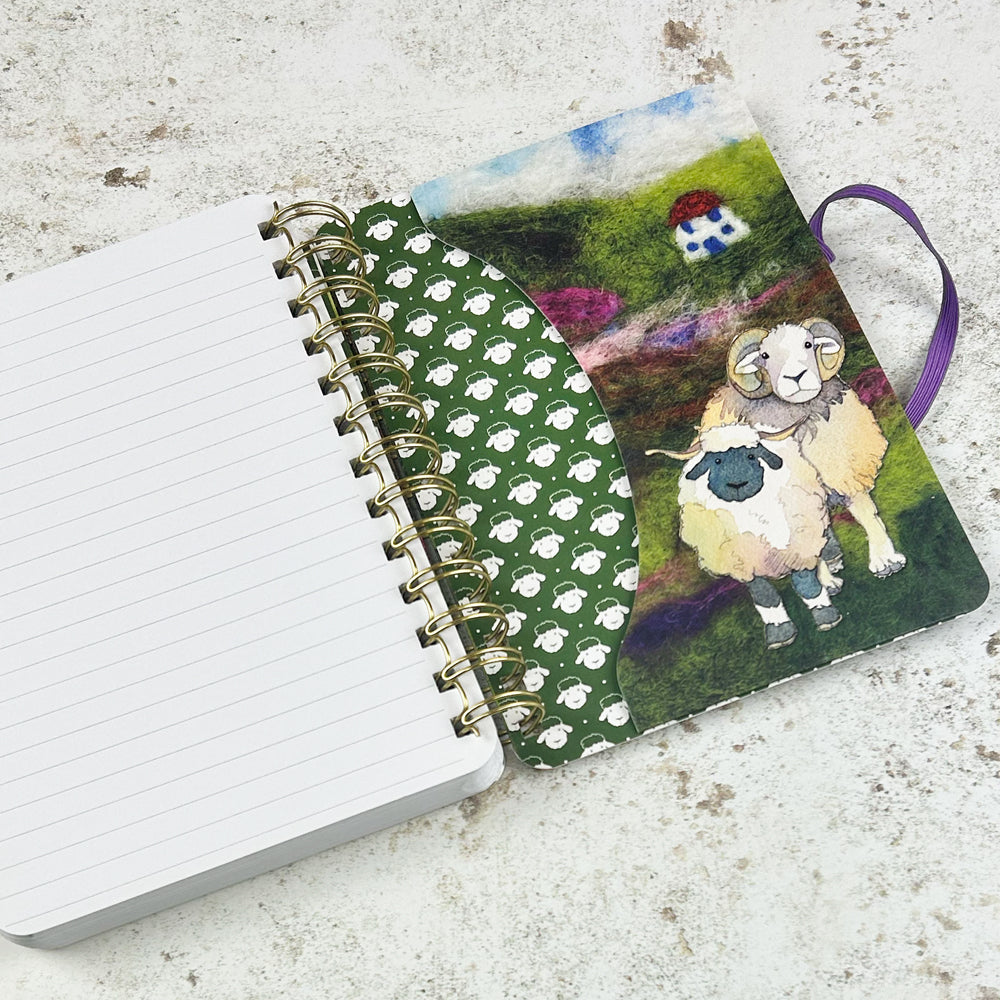 „Felted Sheep" Spiral-Notizbuch von Emma Ball mit lila Gummiband. Filz-Aquarell-Illustration: Schafherde mit Widdern, Mutterschafen und Lämmern auf grünem Hügel, kleines Haus im Hintergrund.