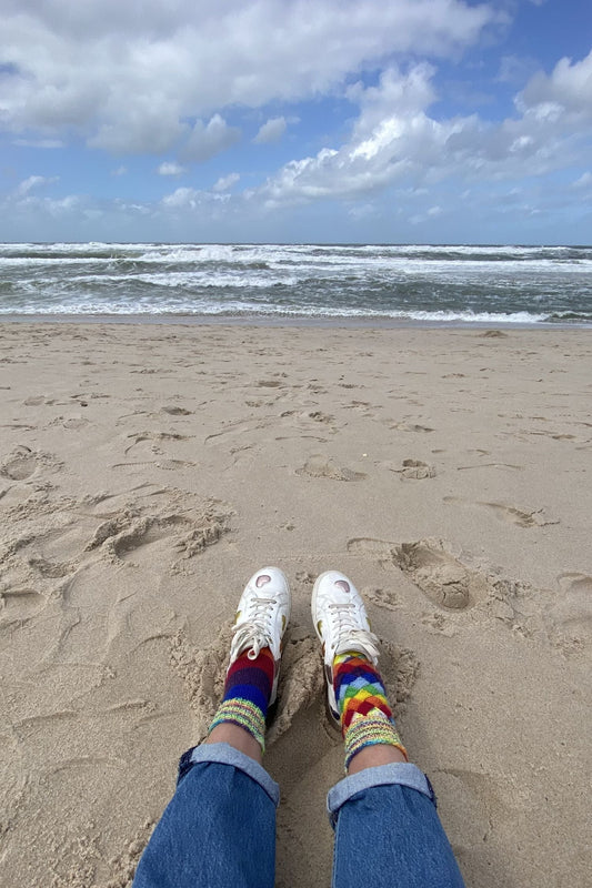 Füße in selbstgestrickten Entrelac-Socken in Regenbogenfarben am Sandstrand mit Blick aufs Meer – die fertige Anleitung in Aktion am Wasser