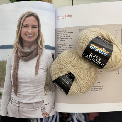 Aufgeschlagenes Buch mit der Seite zum Modell „Rope Mini" — darauf liegt ein cremefarbenes Knäuel Sesia Super Cashmere; links im Buch eine Person mit einem schmalen braunen Strukturhalstuch am Strand.