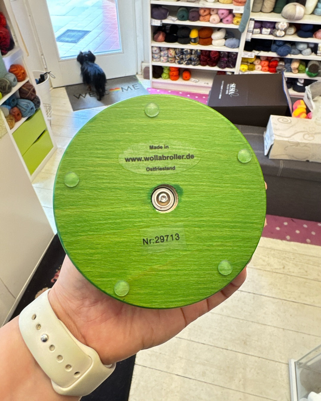 
Unterseite eines grünen Wollabrollers in einer Hand gehalten: sichtbar das integrierte Kugellager in der Mitte, vier Gleitfüße am Rand, Produktionsnummer Nr: 29713 und Herstellerangabe „Made in Ostfriesland, www.wollabroller.de". Im Hintergrund ein Wollladen mit Regalen voller Garnknäuel.