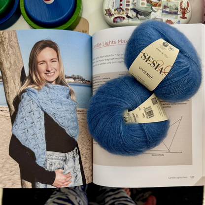 Aufgeschlagenes Buch mit der Seite zum Modell „Candle Lights Maxi" — darauf liegt ein kornblumenblaues Knäuel Sesia Vivienne (Mohair-Mix); links im Buch eine Person mit einem großen blauen Lace-Tuch am Strand.