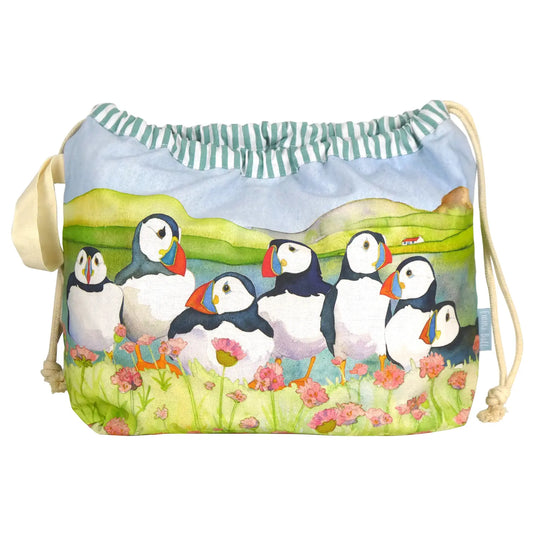 Projektbeutel „Seathrift Puffins" von Emma Ball auf weißem Hintergrund — Baumwollbeutel mit Kordelzug, bedruckt mit sechs Papageientauchern zwischen rosa Strandnelken vor grüner Küstenlandschaft; grün-weiß gestreiftes Innenfutter sichtbar am Rand.