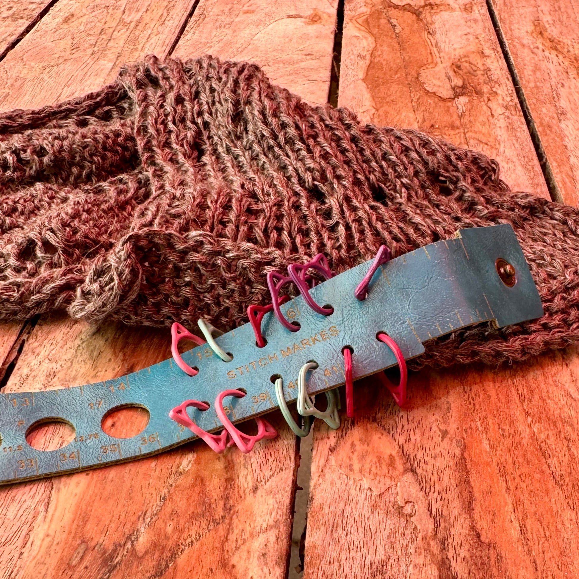 Der Maßgürtel auf einem Holztisch, in die seitlichen Schlitze sind bunte Maschenmarkierer in Katzenform eingehängt. Daneben ein rostrotes Strickstück.