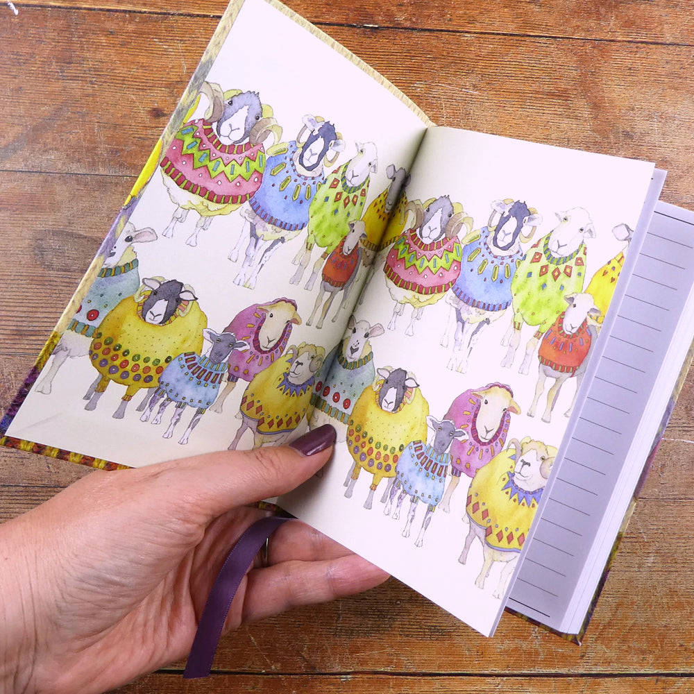 Aufgeklapptes Notizbuch „Sheep in Sweaters" von Emma Ball — bedruckter Innendeckel links zeigt eine bunte Schafherde in gemusterten Pullovern in Aquarell; rechts die ersten linierten Seiten; lila Lesebändchen sichtbar.
