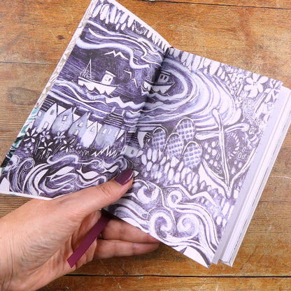 Aufgeschlagenes „Fishing Village" Notizbuch von Emma Ball mit Schwarz-Weiß-Illustration der Innenseiten: Häuser am Hafen, Fischerboote, Wellen und Blumen. Eine Hand hält das Buch über einer Holzfläche.