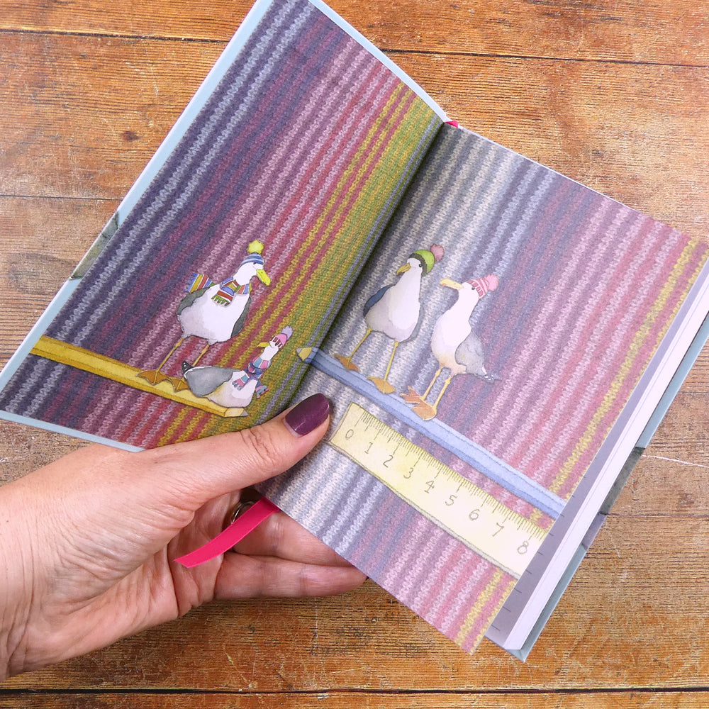Eine Hand hält ein aufgeklapptes Notizbuch „Gulls in Beanies" von Emma Ball — auf dem Einband Möwen in bunten Strickpullovern und Schals auf gestreiftem Strickmuster-Hintergrund; ein Lineal liegt quer über dem Buch.