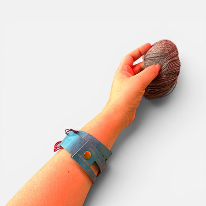 Der Maßgürtel ums Handgelenk geschlossen, mit Druckknopf. Sichtbar sind die Skala in Zoll und Zentimeter und das Zählfenster. Die Hand greift nach einem Wollknäuel in gedämpftem Rotbraun.