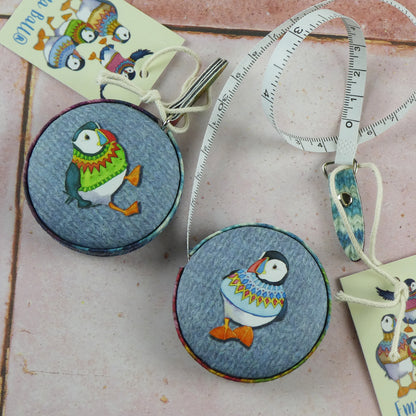 Zwei „Woolly Puffins“ Maßbänder von Emma Ball auf rosafarbenem Fliesenhintergrund — runde Gehäuse mit Papageientauchern in bunt gemusterten Strickpullovern; eines mit ausgezogenem weißem Maßband mit Zentimeter- und Zollskala, Emma-Ball-Anhänger daneben.