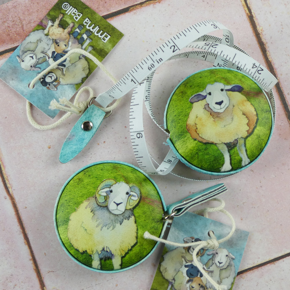 Zwei „Felted Sheep“ Maßbänder von Emma Ball auf rosafarbenem Fliesenhintergrund — eines mit Schafmotiv und ausgezogenem weißem Maßband, das zweite mit Widdermotiv; Anhänger mit weiteren Schafillustrationen daneben.
