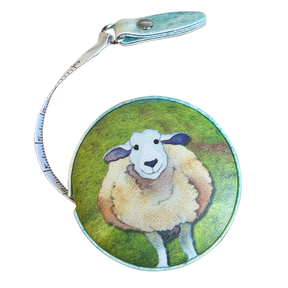 Geöffnetes „Felted Sheep“ Maßband von Emma Ball auf weißem Hintergrund — rundes Gehäuse mit wolligem Schaf auf grünem Hintergrund, teilweise ausgezogenes weißes Maßband und türkisfarbene Lasche mit Druckknopf.