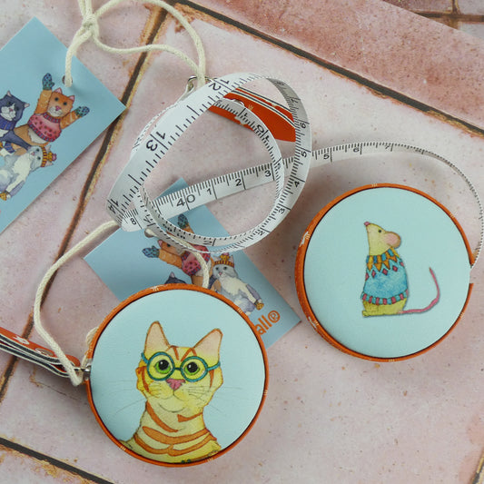 Zwei „Kittens in Mittens" Maßbänder von Emma Ball auf rosa Fliesen: orange Katze mit grüner Brille und Maus im Strickpullover. Daneben Anhänger mit Katzenmotiv und ausgezogenes Maßband mit Zentimeter- und Inch-Skala.