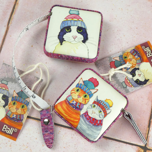 Zwei „Cats in Hats" Maßbänder von Emma Ball auf rosa Fliesen. Vorder- und Rückseite mit Aquarell-Illustrationen verschiedener Katzen in Strickmützen, dazu Anhänger mit Katzenmotiven.