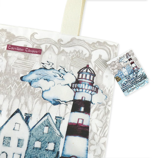 Nahaufnahme der „Fishing Village" Tragetasche von Caroline Cleave — Leuchtturm, Hafenhäuser und Möwen in Blau- und Grautönen im Linogravüre-Stil; Caroline Cleave-Label und bedrucktes Anhänger-Etikett erkennbar.