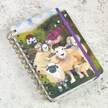 „Felted Sheep" Spiral-Notizbuch von Emma Ball mit lila Gummiband. Filz-Aquarell-Illustration: Schafherde mit Widdern, Mutterschafen und Lämmern auf grünem Hügel, kleines Haus im Hintergrund.