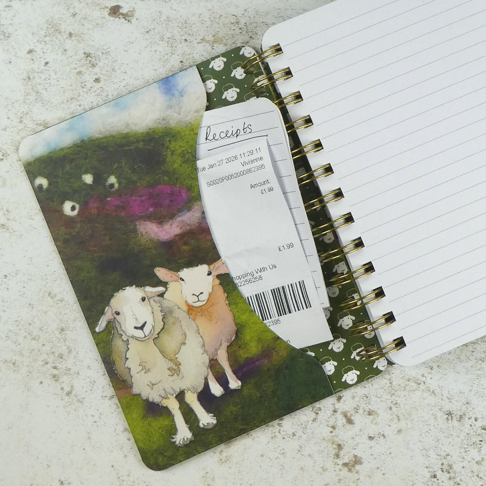 
Innentasche des „Felted Sheep" Notizbuchs mit einer eingesteckten Quittung. Filz-Aquarell-Illustration von Emma Ball: zwei Schafe auf grünem Hügel, Muster aus kleinen Schafsköpfen.