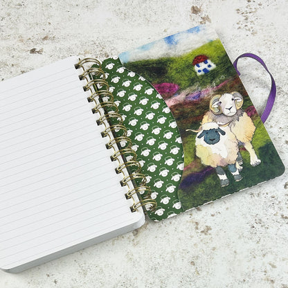 
„Felted Sheep" Spiral-Notizbuch von Emma Ball mit lila Gummiband. Filz-Aquarell-Illustration: Schafherde mit Widdern, Mutterschafen und Lämmern auf grünem Hügel, kleines Haus im Hintergrund.