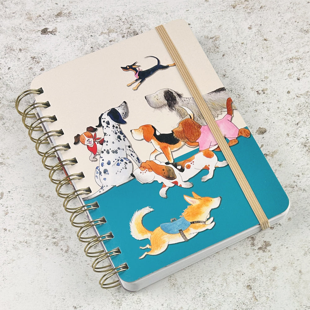 „Paw Club" Spiral-Notizbuch von Eric Heyman mit beigem Gummiband. Cremefarbene obere Hälfte mit Aquarell-Illustration: Dalmatiner, Beagle, Bassets, Spaniel, kleiner Hund mit rotem Halstuch. Untere Hälfte in Petrolblau mit Corgi und springendem Hund.
