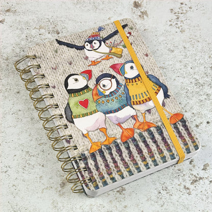 Geschlossenes Spiral-Notizbuch „Woolly Puffin" von Emma Ball mit goldgelbem Gummiband — Einband zeigt vier Papageientaucher in selbst gestrickten Pullovern auf cremefarbenem Hintergrund mit Strickmuster-Bordüre unten.