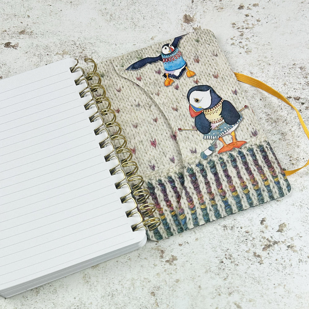 Aufgeschlagenes Spiral-Notizbuch „Woolly Puffin" von Emma Ball — linke Seite leer und liniert, rechte Seite zeigt den Einband mit zwei strickenden Papageientauchern in bunten Pullovern auf cremefarbenem Strickmuster-Hintergrund.