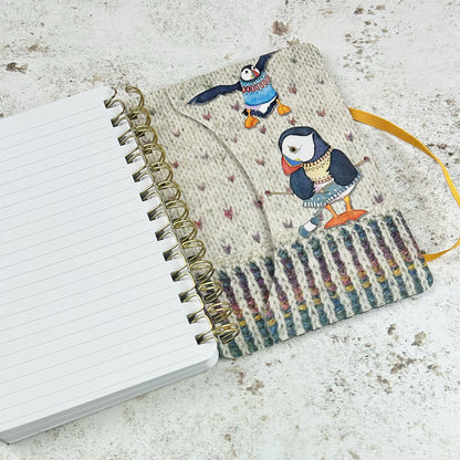 Aufgeschlagenes Spiral-Notizbuch „Woolly Puffin" von Emma Ball — linke Seite leer und liniert, rechte Seite zeigt den Einband mit zwei strickenden Papageientauchern in bunten Pullovern auf cremefarbenem Strickmuster-Hintergrund.
