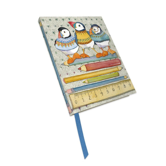 „Woolly Puffin" Notizbuch von Emma Ball mit gebundenem Einband. Aquarell-Illustration: drei Papageientaucher in bunten Strickpullovern auf einer Reihe Buntstifte und einem Lineal. Mit blauem Lesezeichenband.