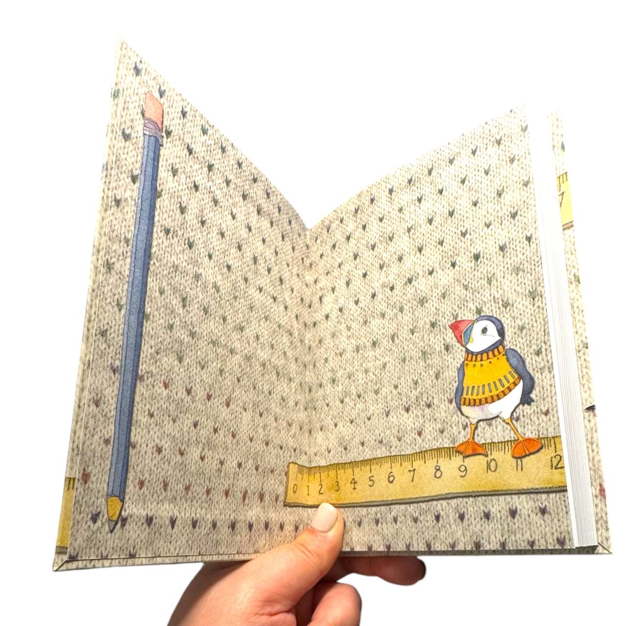 Aufgeschlagenes „Woolly Puffin" Notizbuch von Emma Ball mit bedrucktem Innenumschlag: Strickmuster-Hintergrund, Bleistift, Lineal und ein Papageientaucher im gelben Strickpullover. Eine Hand hält das Buch.