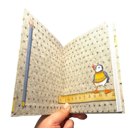 Aufgeschlagenes „Woolly Puffin" Notizbuch von Emma Ball mit bedrucktem Innenumschlag: Strickmuster-Hintergrund, Bleistift, Lineal und ein Papageientaucher im gelben Strickpullover. Eine Hand hält das Buch.