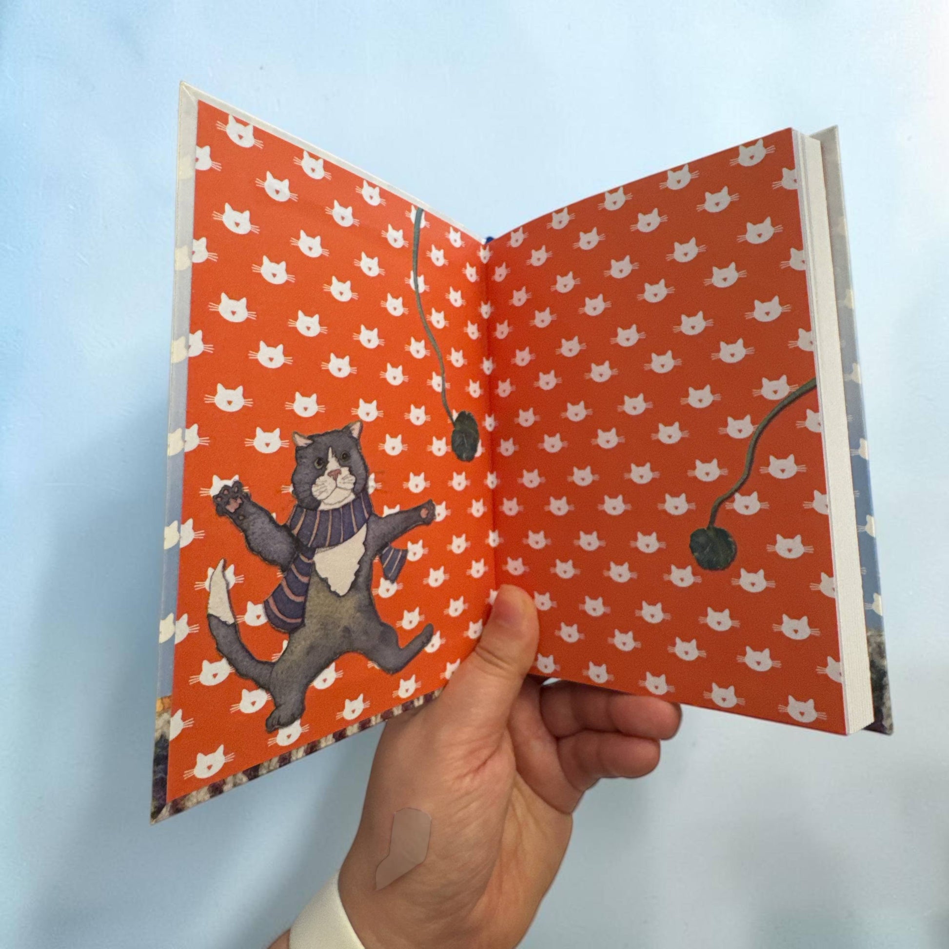 Aufgeklapptes Notizbuch „Kittens in Mittens" — bedruckte Innendeckel in leuchtendem Rotorange mit weißem Katzenmuster; links ein tanzender grauer Kater im Schal, rechts das Lesebändchen sichtbar.