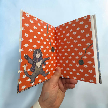 Aufgeklapptes Notizbuch „Kittens in Mittens" — bedruckte Innendeckel in leuchtendem Rotorange mit weißem Katzenmuster; links ein tanzender grauer Kater im Schal, rechts das Lesebändchen sichtbar.