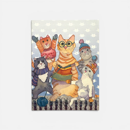 Vorderseite des Notizbuchs „Kittens in Mittens" von Emma Ball — fünf Katzen in Strickpullovern, Mützen und Fäustlingen auf hellblauem Hintergrund mit Katzenmuster; die mittlere Katze trägt Brille und strickt, unten ein blaues Wollknäuel; Strickmuster-Bordüre am unteren Rand.